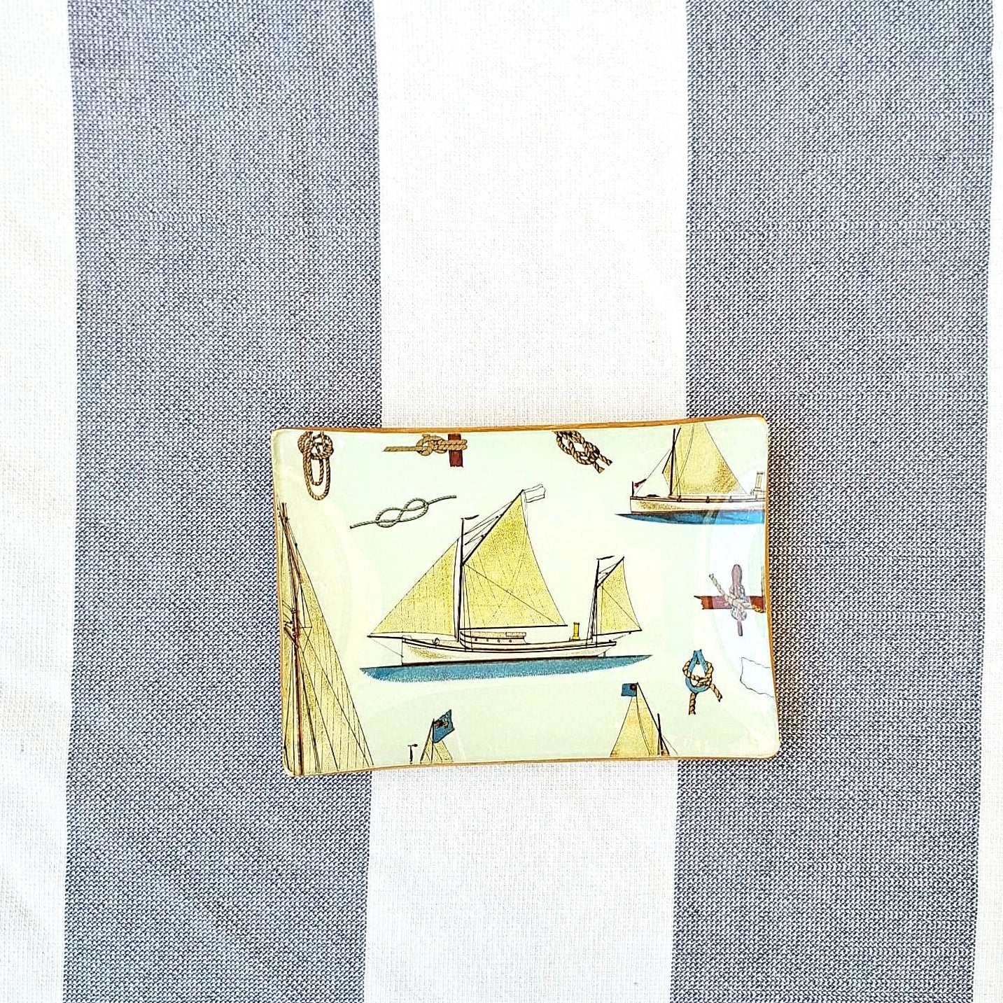 Bandeja 16x11 cm decorada a mano con estampado náutico de veleros sobre fondo beige. El diseño presenta una embarcacion de vela en tonos beige, marrón y azul. El patrón incluye también elementos marinos como anclas, nudos marineros y pequeños detalles náuticos distribuidos entre las embarcaciones. Las bandejas tienen bordes con ribete dorado. Artesanía Española Dmarta