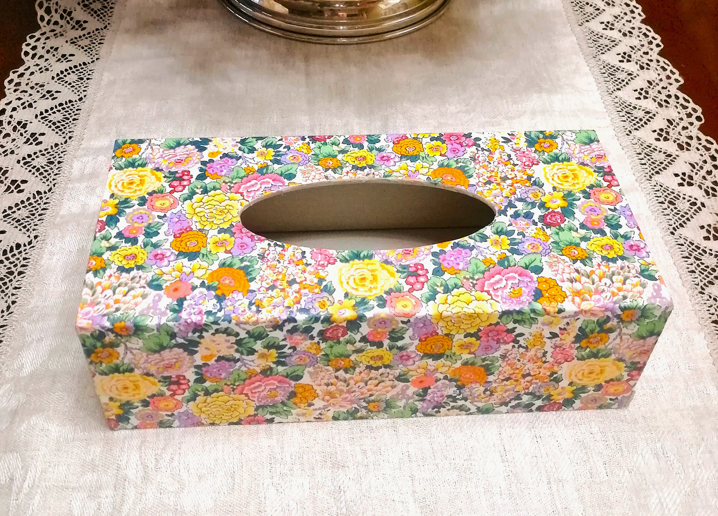 "Caja dispensadora de pañuelos decorada a mano con estampado floral Liberty. El diseño presenta flores pequeñas en tonos pastel incluyendo rosa, amarillo, morado, verde y naranja sobre fondo blanco. Es ideal para decoración del hogar, baños, dormitorios o como regalo funcional." Realizada en España