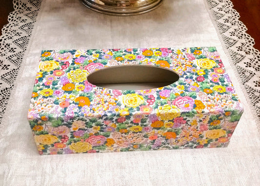 "Caja dispensadora de pañuelos decorada a mano con estampado floral Liberty. El diseño presenta flores pequeñas en tonos pastel incluyendo rosa, amarillo, morado, verde y naranja sobre fondo blanco. Es ideal para decoración del hogar, baños, dormitorios o como regalo funcional." Realizada en España
