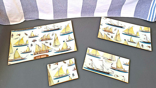 Conjunto de bandejas decoradas a mano con estampado náutico de veleros sobre fondo beige. El diseño presenta una variedad de embarcaciones de vela en tonos beige, marrón y azul, incluyendo veleros clásicos, barcos de pesca tradicionales y yates con diferentes tipos de velas. El patrón incluye también elementos marinos como anclas, nudos marineros y pequeños detalles náuticos distribuidos entre las embarcaciones. Las bandejas tienen bordes con ribete dorado y están disponibles en varios tamaños rectangulares