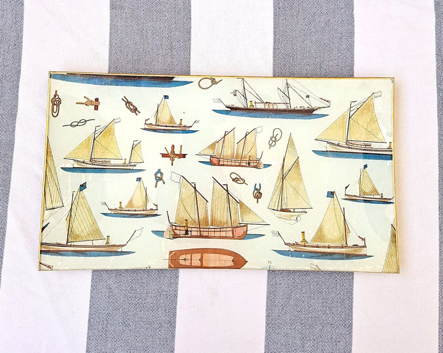 Bandejas decorada a mano con estampado náutico de veleros sobre fondo beige. El diseño presenta una variedad de embarcaciones de vela en tonos beige, marrón y azul, incluyendo veleros clásicos, barcos de pesca tradicionales y yates con diferentes tipos de velas. El patrón incluye también elementos marinos como anclas, nudos marineros y pequeños detalles náuticos distribuidos entre las embarcaciones. Las bandejas tienen bordes con ribete dorado. Artesanía española Dmarta.