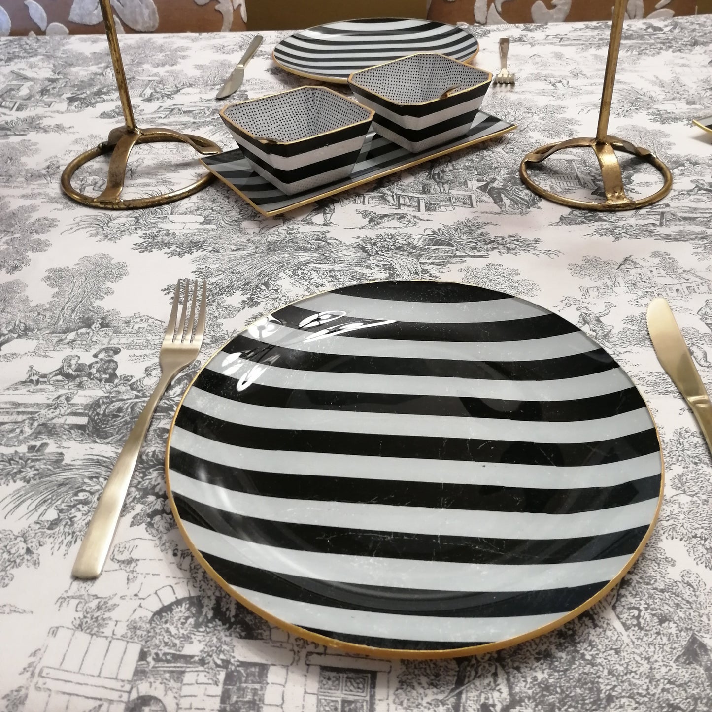 "Mesa con vajilla de cristal decorada a mano de diseño contemporáneo con patrón de rayas negras sobre fondo blanco, candelabros ornamentales dorados con motivo de palmeras, cubiertos dorados y mantel estilo toile con escenas clásicas en tonos grises"
✅ Decoración 100% artesanal
✅ Diseño atemporal y elegante
✅ Perfecta para ocasiones especiales
✅ Combina con cualquier estilo decorativo
✅ Piezas únicas e irrepetibles