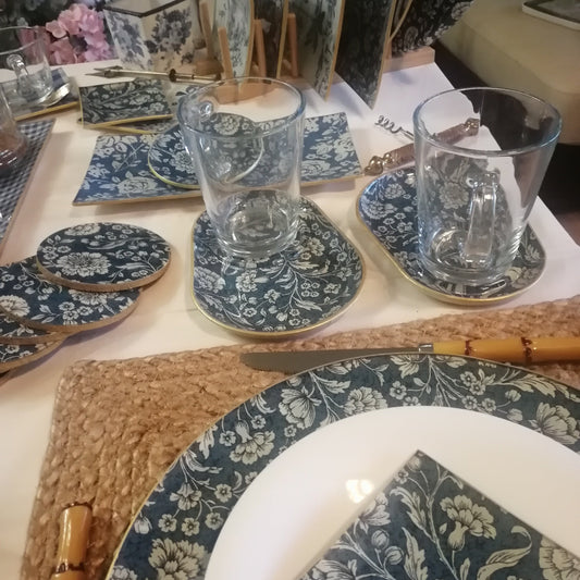 Detalle de productos artesanales decoupage cristal con estampado floral en crudo sobre fondo azul: Bajoplato 30cm, tazas desayuno, posavasos, bandejas aperitivos y platito pan. 