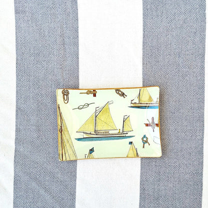  Bandeja 16x11 cm decorada a mano con estampado náutico de veleros sobre fondo beige. El diseño presenta una  embarcacion de vela en tonos beige, marrón y azul. El patrón incluye también elementos marinos como anclas, nudos marineros y pequeños detalles náuticos distribuidos entre las embarcaciones. Las bandejas tienen bordes con ribete dorado. Artesanía Española Dmarta