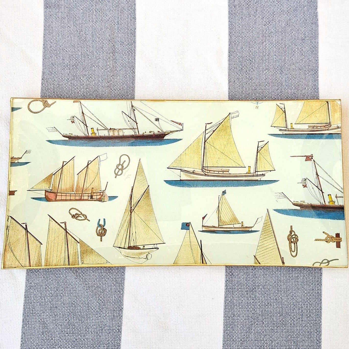 Bandejas decorada a mano con estampado náutico de veleros sobre fondo beige. El diseño presenta una variedad de embarcaciones de vela en tonos beige, marrón y azul, incluyendo veleros clásicos, barcos de pesca tradicionales y yates con diferentes tipos de velas. El patrón incluye también elementos marinos como anclas, nudos marineros y pequeños detalles náuticos distribuidos entre las embarcaciones. Las bandejas tienen bordes con ribete dorado. Artesanía española Dmarta