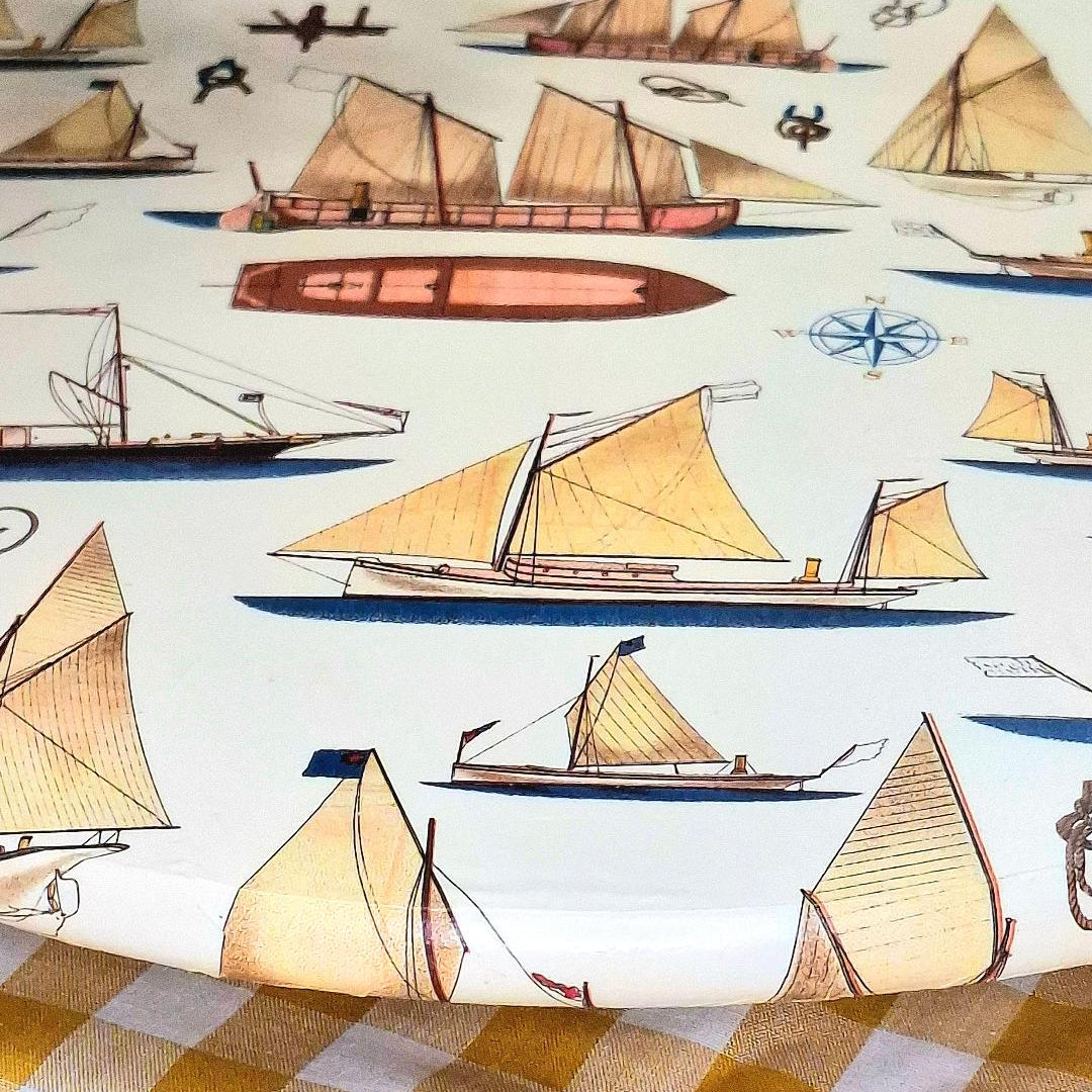"Detalle de la tabla giratoria Marina Veleros con ilustraciones de barcos y veleros náuticos en tonos beige, azul y terracota sobre mantel de cuadros vichy amarillo"