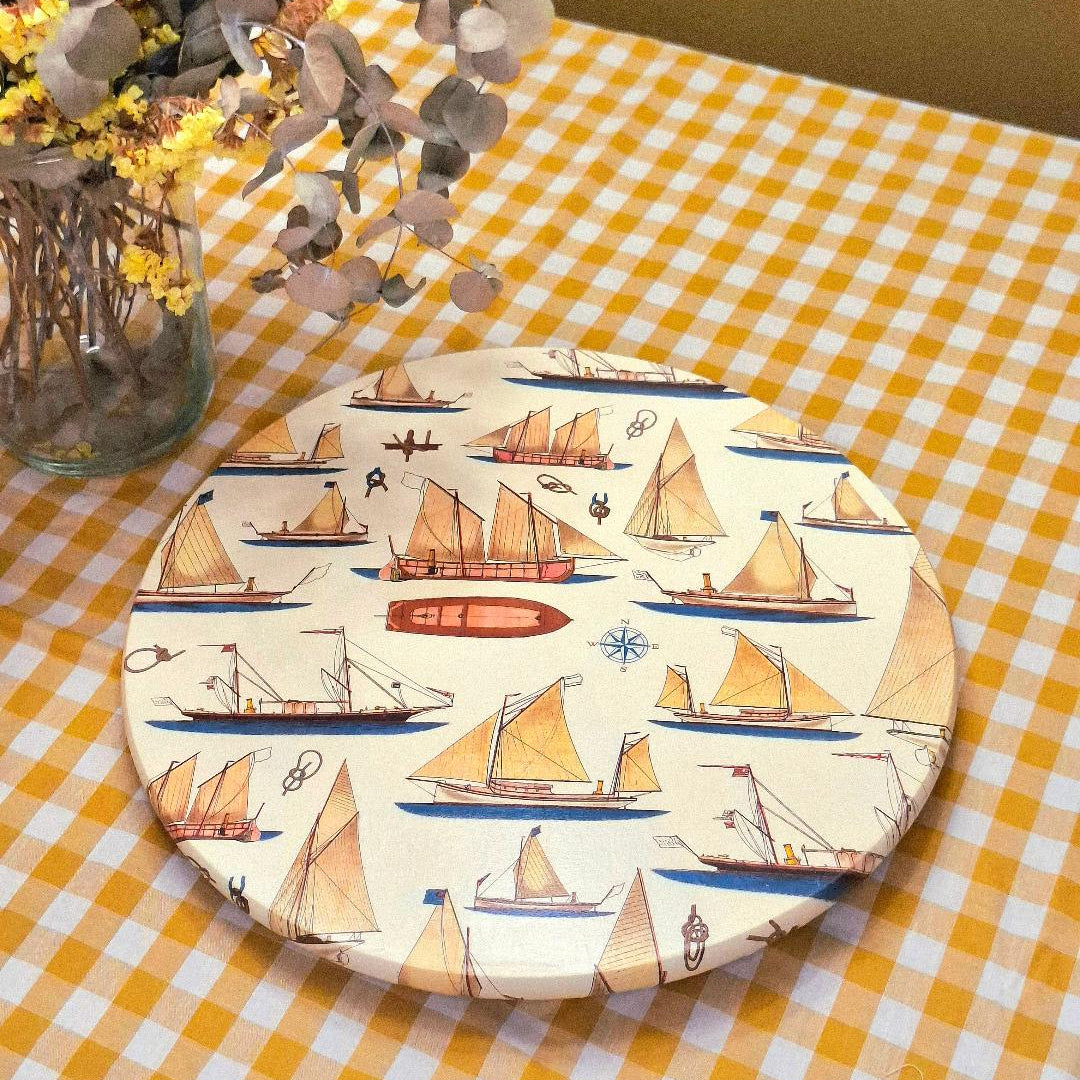 "Tabla giratoria Marina Veleros con diseño de barcos náuticos sobre mantel de cuadros vichy amarillo y blanco, junto a florero con flores secas"