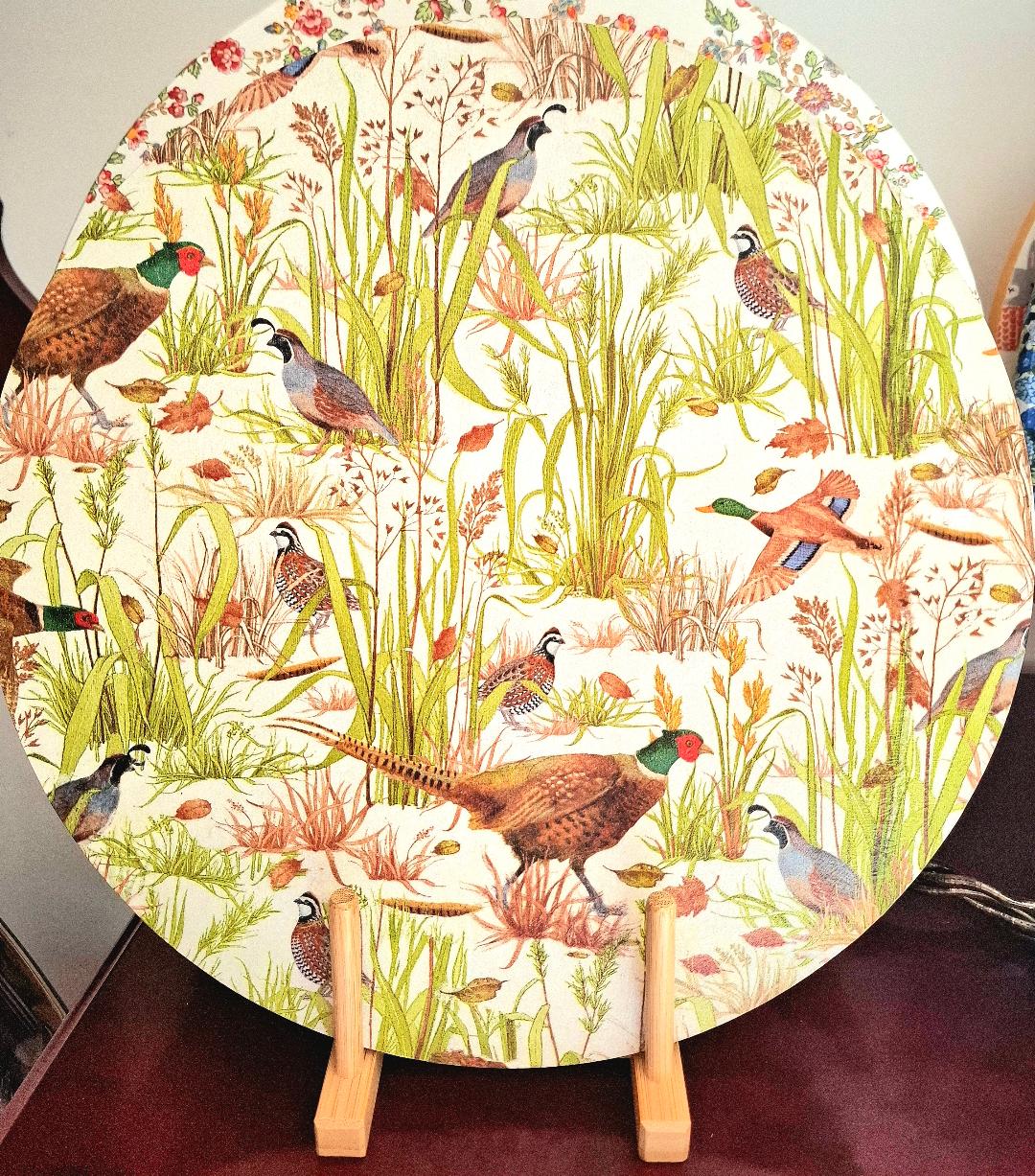 "Tabla giratoria decorativa con diseño de faisanes y aves de caza entre vegetación silvestre y flores en tonos tierra, verde y naranja, presentada sobre soporte de madera"
