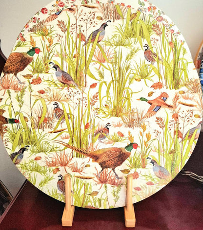 "Tabla giratoria decorativa con diseño de faisanes y aves de caza entre vegetación silvestre y flores en tonos tierra, verde y naranja, presentada sobre soporte de madera"
